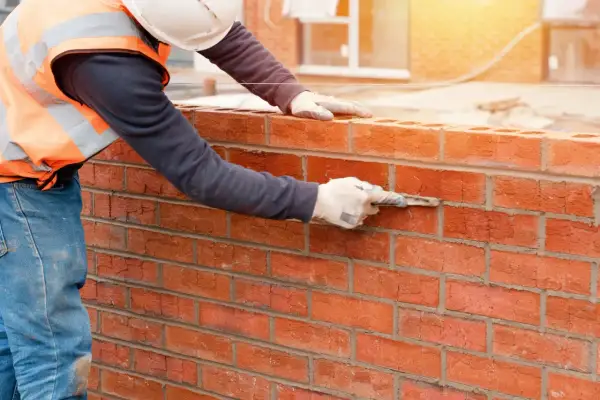 Muratore con guanti e cazzuola stende malta modellabile tra i mattoni rossi di un muro in costruzione.
