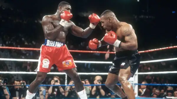 Two boxers, one in red shorts with "BRUNO" on the waistband, the other in black shorts, face off in a tense boxing match. The pressure fighter in red throws a punch.