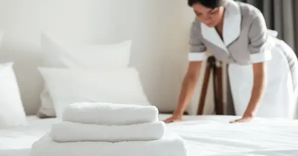 Pokojówka w hotelu dba o czystość i porządek. Na pierwszym planie stos białych ręczników, w tle kobieta poprawia pościel.