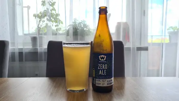Szklanka i butelka piwa bezalkoholowego "Zero Ale" na drewnianym stole. Etykieta informuje, co zawiera piwo bezalkoholowe: ekstrakt 6%, alkohol ≤0.5%.