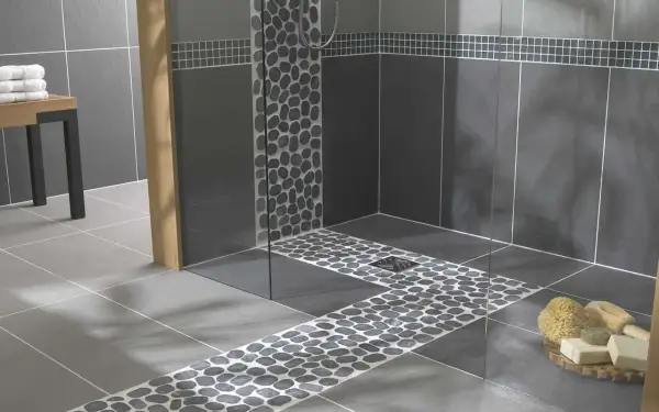 Douche italienne moderne avec carrelage imitation pierre de galets sur le sol et les murs. Un banc en bois et des serviettes ajoutent une touche chaleureuse.