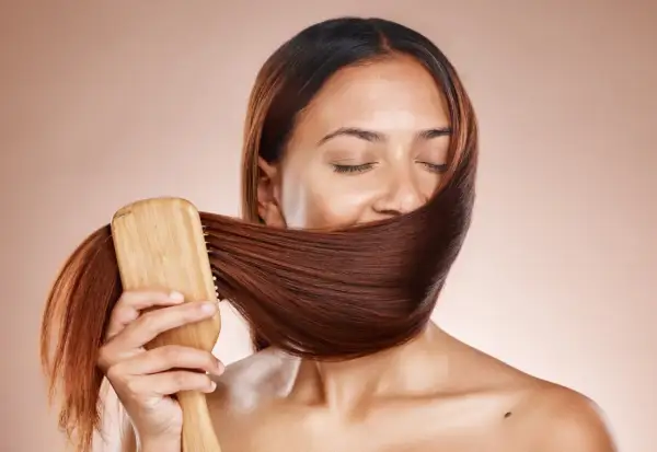 Frauen mit geschlossenen Augen bürsten ihr glänzendes, braunes Haar mit einer Holzbürste. Ein Moment der Ruhe und des **Haare pflegen**.