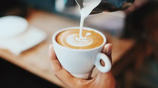 En hand håller en vit kopp med en vacker kaffe latte, där mjölken skapar ett hjärta i skummet.