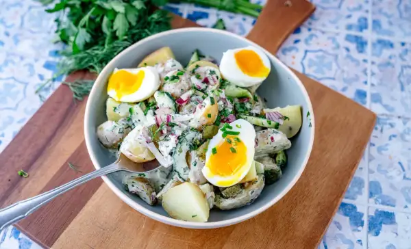 Schüssel mit cremigem Kartoffelsalat mit Gurken, Zwiebeln und gekochten Eiern, garniert mit frischen Kräutern.
