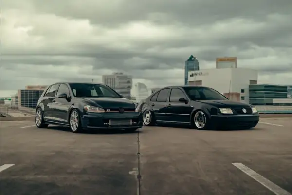 Dwa czarne Volkswageny: sportowy hatchback GTI i obniżony sedan, stoją na parkingu z widokiem na miasto.