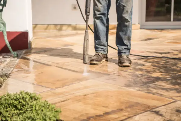 Un homme nettoie sa terrasse carrelage avec un nettoyeur haute pression. L'eau éclabousse les dalles.