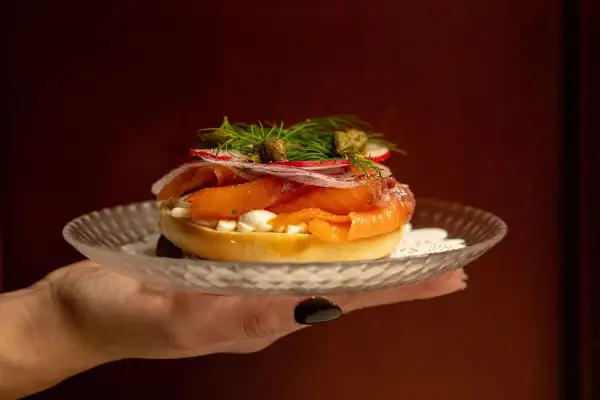 En hand håller upp en bagel med rökt lax, färskost, rödlök, rädisor och kapris. Perfekt för en lyxig bank hotel middag.