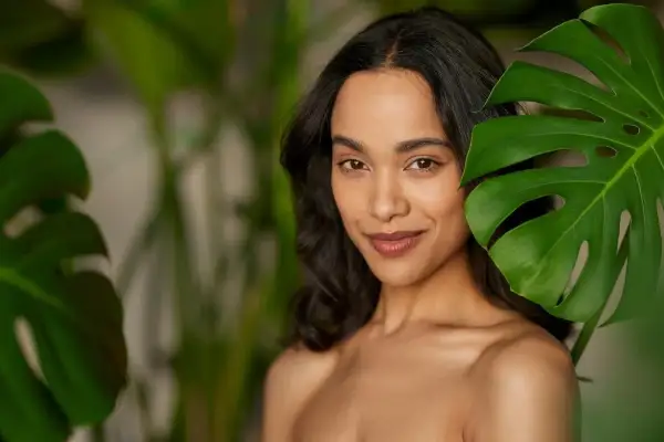 Mujer con maquillaje efecto bronceado, sonriendo entre hojas verdes.