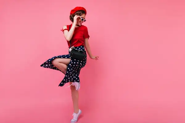 Urocza dziewczyna w stylu pin-up girl w czerwonej bluzce, spódnicy w grochy i czerwonym beretcie, skacze z radości na różowym tle.