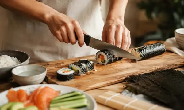 Ręce kroją rolkę sushi na desce. Widać składniki: ryż, łososia, ogórka. Pyszne domowe sushi w przygotowaniu.