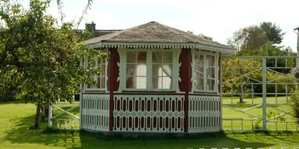 Pavillon de jardin rouge et blanc, orné de dentelle de bois. Une pomme tombe d'un arbre, peut-être pour échapper à la taxe abri de jardin.