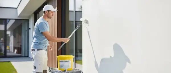 Mann streicht mit einer Fassadenrolle eine weiße Wand. Ein Eimer mit Farbe steht daneben.