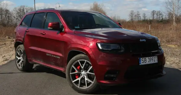 Czerwony Jeep Grand Cherokee SRT z czarnymi felgami i czerwonymi zaciskami hamulcowymi, zaparkowany na drodze wśród suchych traw i drzew.