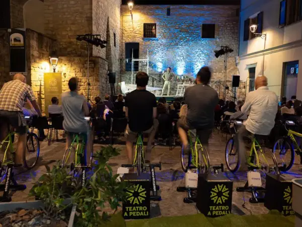 Spettatori pedalano su cyclette per alimentare un'esibizione di teatro a pedali in una piazza illuminata.