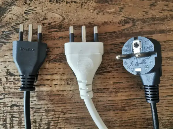 Trzy wtyczki elektryczne, w tym dwie z trzema bolcami, typowe dla gniazdek we Włoszech, leżą na drewnianym blacie.