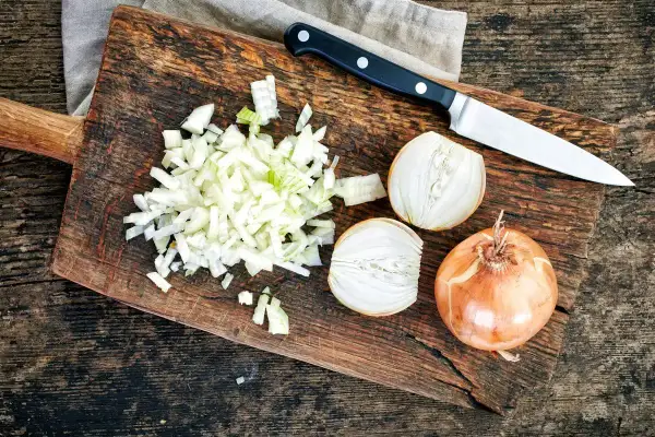 Cipolla cruda digeribile, tagliata finemente su un tagliere di legno, accanto a una cipolla intera e a metà. Un coltello è pronto all'uso.