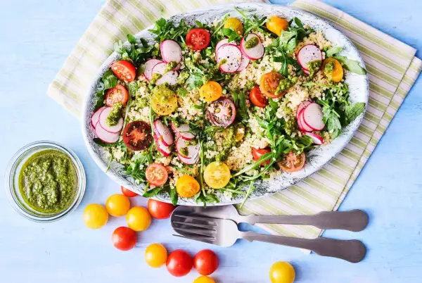 En stor bulgursallad med körsbärstomater, rädisor och ruccola, toppad med en grön dressing. Serveras med en skål pesto och extra tomater.