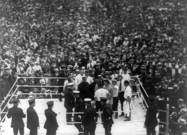Boxing ring with fighters and officials, surrounded by a massive, cheering crowd in vintage attire.