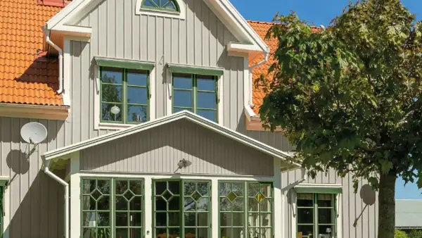 Vitt hus med gråa knutar och orange tak. Gröna fönster och en stor lönn skänker skugga.