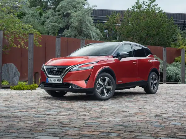 Czerwony Nissan Qashqai 2025 z czarnym dachem, zaparkowany na brukowanej nawierzchni, otoczony zielenią i industrialnym ogrodzeniem.