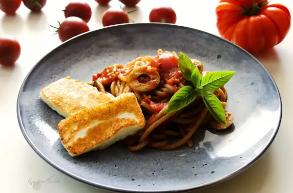 Makaron z halloumi na talerzu, z sosem pomidorowym, oliwkami i bazylią. Obok leżą świeże pomidory.