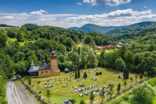 Drewniana cerkiew i cmentarz w malowniczej dolinie Beskidu Niskiego, otoczone bujną zielenią lasów i wzgórz.