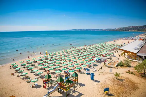 Słoneczna plaża w Złote Piaski pełna leżaków i parasoli, z ludźmi kąpiącymi się w morzu i bawiącymi na placu zabaw.