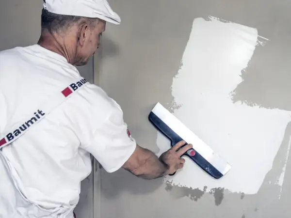 Fachowiec w białej czapce i koszulce z logo Baumit wykonuje gipsowanie ścian, używając szerokiej pacy do rozprowadzania masy.