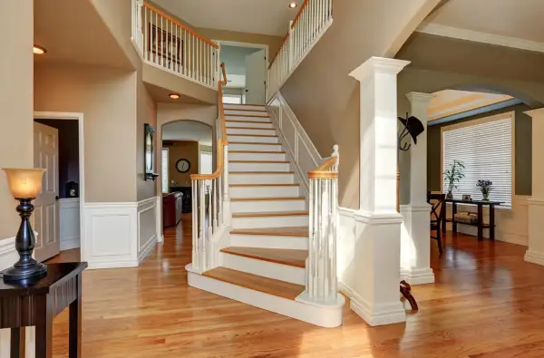 A grand foyer with a sweeping staircase featuring white painted risers and wooden treads. The staircase leads to an upper landing, with a view into a living area.