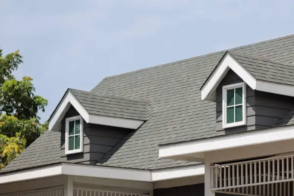 A close-up of a house roof showcasing different types of shingles in a grey hue, with dormer windows peeking out.