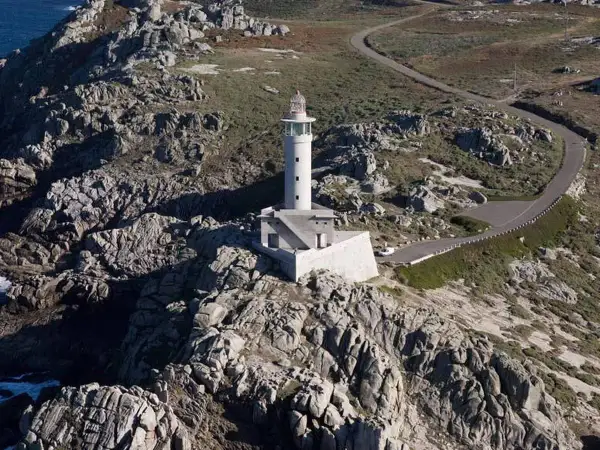 Faro blanco en una punta rocosa, con una carretera serpenteante que sube hacia él.