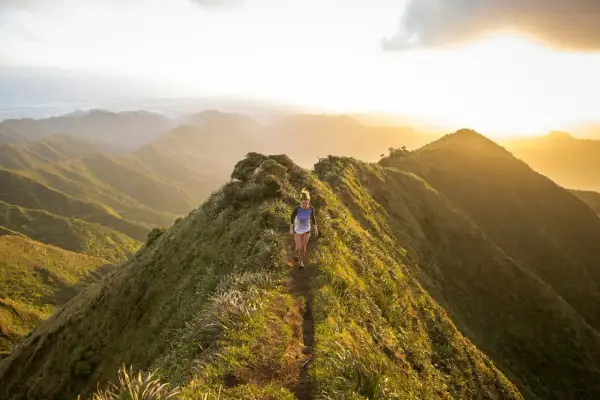 Une femme court sur un sentier de montagne verdoyant au coucher du soleil, profitant d'un parcours spectaculaire.