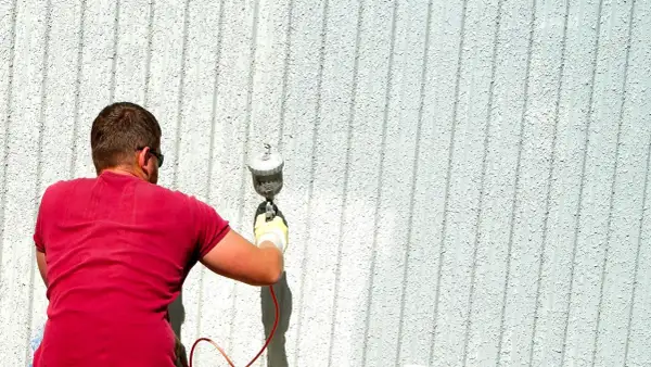 Uomo con maglietta rossa usa uno spruzzatore per verniciare una parete esterna. Ottimi trucchi per verniciare a spruzzo per un lavoro uniforme.