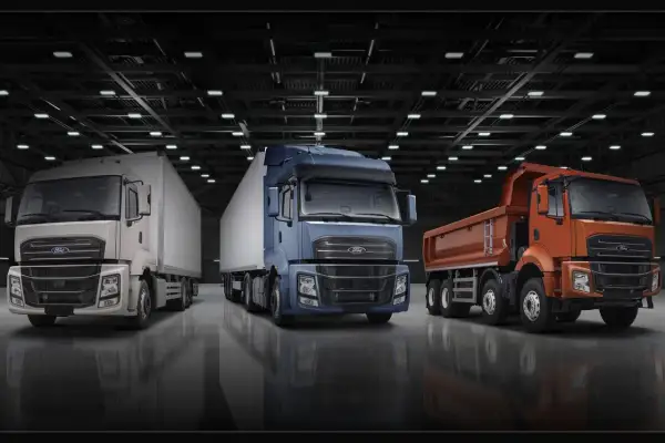 Trois camions Ford F-Line : un blanc avec remorque, un bleu avec remorque, et un orange benne basculante, alignés dans un entrepôt.
