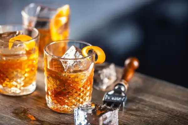 Tre old fashioned-drinkar med is och apelsinskal på ett träbord. En cocktailgaffel ligger bredvid.
