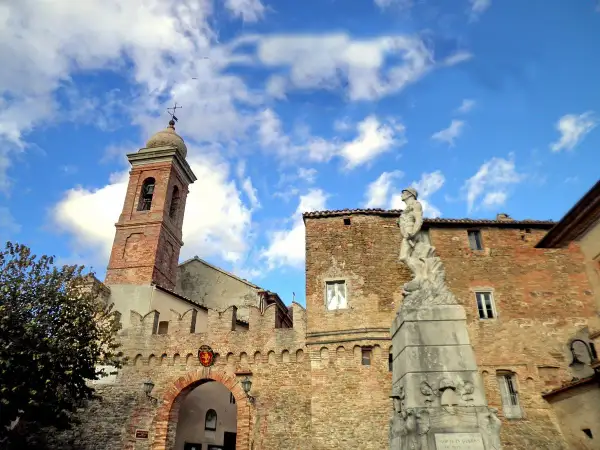 Poggio San Marcello: campanile in mattoni, un arco fortificato e una statua di bronzo sotto un cielo azzurro con nuvole bianche.