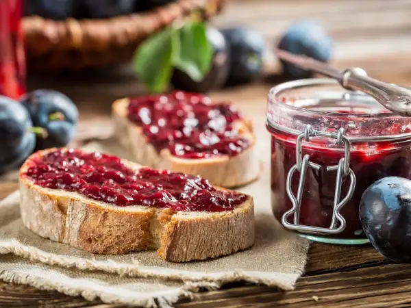 Zwei Scheiben Brot mit Zwetschgenmarmelade mit Zimt, daneben ein Glas der Marmelade und frische Zwetschgen.