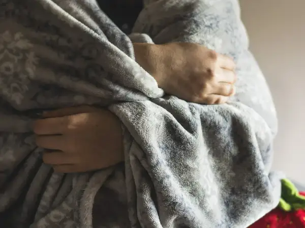 Dłonie skrzyżowane na piersi, otulone miękkim kocem, symbolizują ciągłe uczucie zimna i potrzebę ciepła.