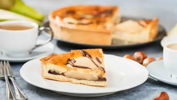 Une part de tarte poire chocolat, garnie de fruits et de copeaux de chocolat, est servie avec une tasse de café et des noisettes.