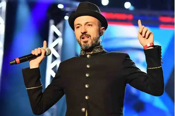 Un cantante con cappello e giacca nera, microfono in mano, evoca le canzoni dei Subsonica con un gesto energico.
