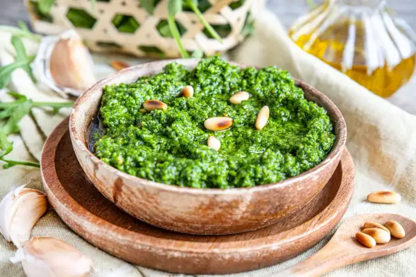 Frisches Rucola Pesto in Holzschale, garniert mit Pinienkernen. Daneben Knoblauch und Olivenöl.