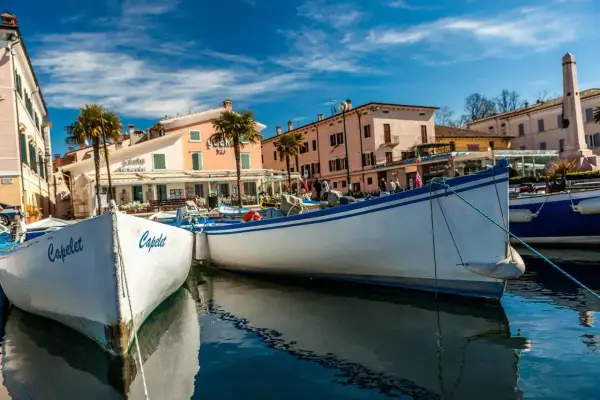 Barche a Cisano di Bardolino, con palme e case colorate sullo sfondo. Il sole splende su un cielo azzurro e limpido.