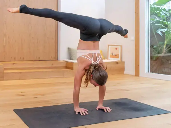 Mujer en postura del arbol yoga, con piernas extendidas en el aire, sobre una esterilla en un estudio luminoso.