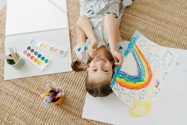Niña sonriente haciendo un arcoíris, rodeada de acuarelas y rotuladores. Perfectas actividades de verano para niños creativos.