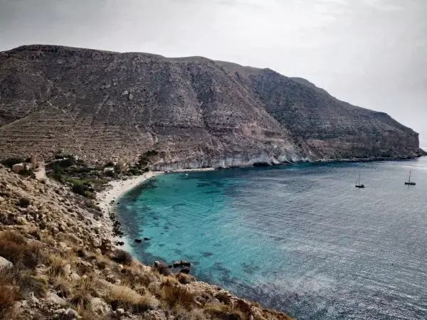 Bahía de aguas turquesas en la playa Cabo de Gata, con barcos navegando y montañas áridas al fondo.