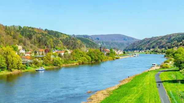 Blick auf den Elberadweg mit Radfahrern und einer Stadt am Flussufer. Die Elbe fließt durch eine grüne Landschaft mit bewaldeten Hügeln. Die Elberadweg Entfernungen sind hier gut zu erkunden.