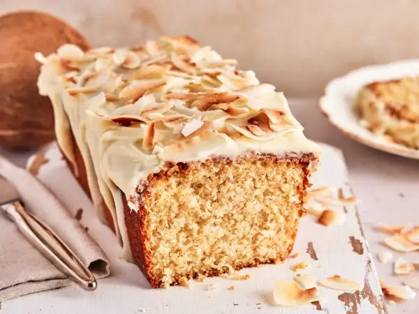 Ein saftiger Kokos Kuchen mit weißer Glasur und gerösteten Kokoschips. Daneben liegt eine Kokosnuss.