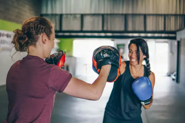 Due donne si allenano nel **boxing** in palestra. Una tiene i guantoni da allenamento, l'altra indossa un guantone blu.