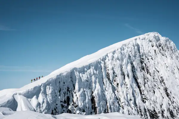 En grupp vandrare på en snötäckt bergstopp under en helags topptur. Den isiga bergssidan kontrasterar mot den klara blå himlen.