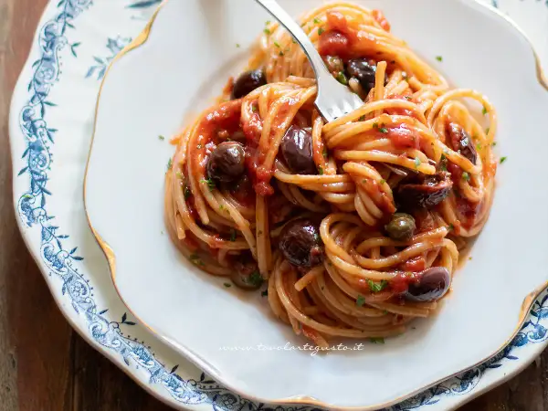 Un piatto invitante di spaghetti alla puttanesca, con olive nere, capperi e un sugo rosso vivace, pronto per essere gustato.